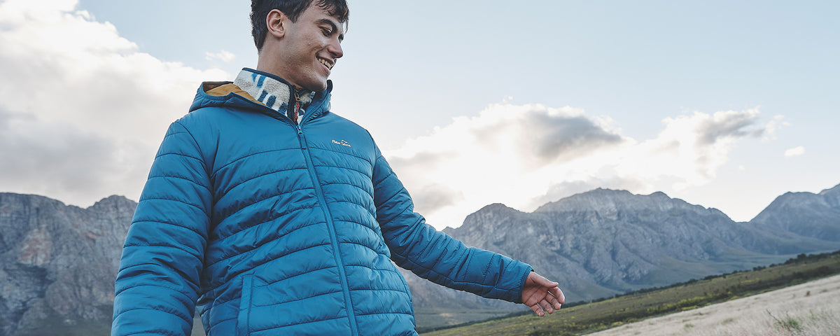 A man wearing a bright blue puffer jacket stands outdoors in front of mountains under a cloudy sky, smiling and looking to the side.